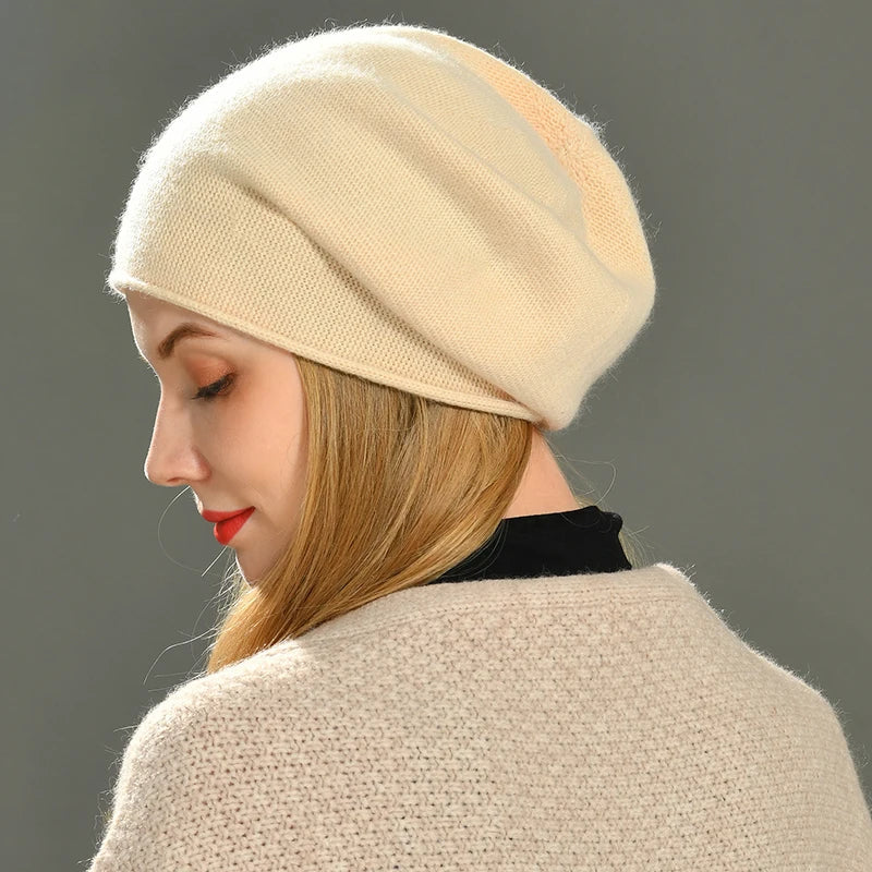 A woman with long blonde hair wears a cream-colored, slouchy cashmere beanie and a matching sweater. She is looking down with her eyes closed, shown from the side against a plain gray background.