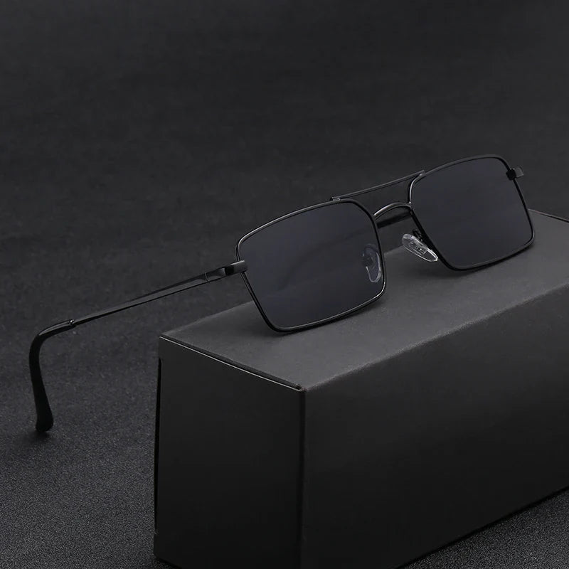 A pair of black rectangular sunglasses with thin, lightweight alloy frames rests on a black box, set against a dark, textured background.
