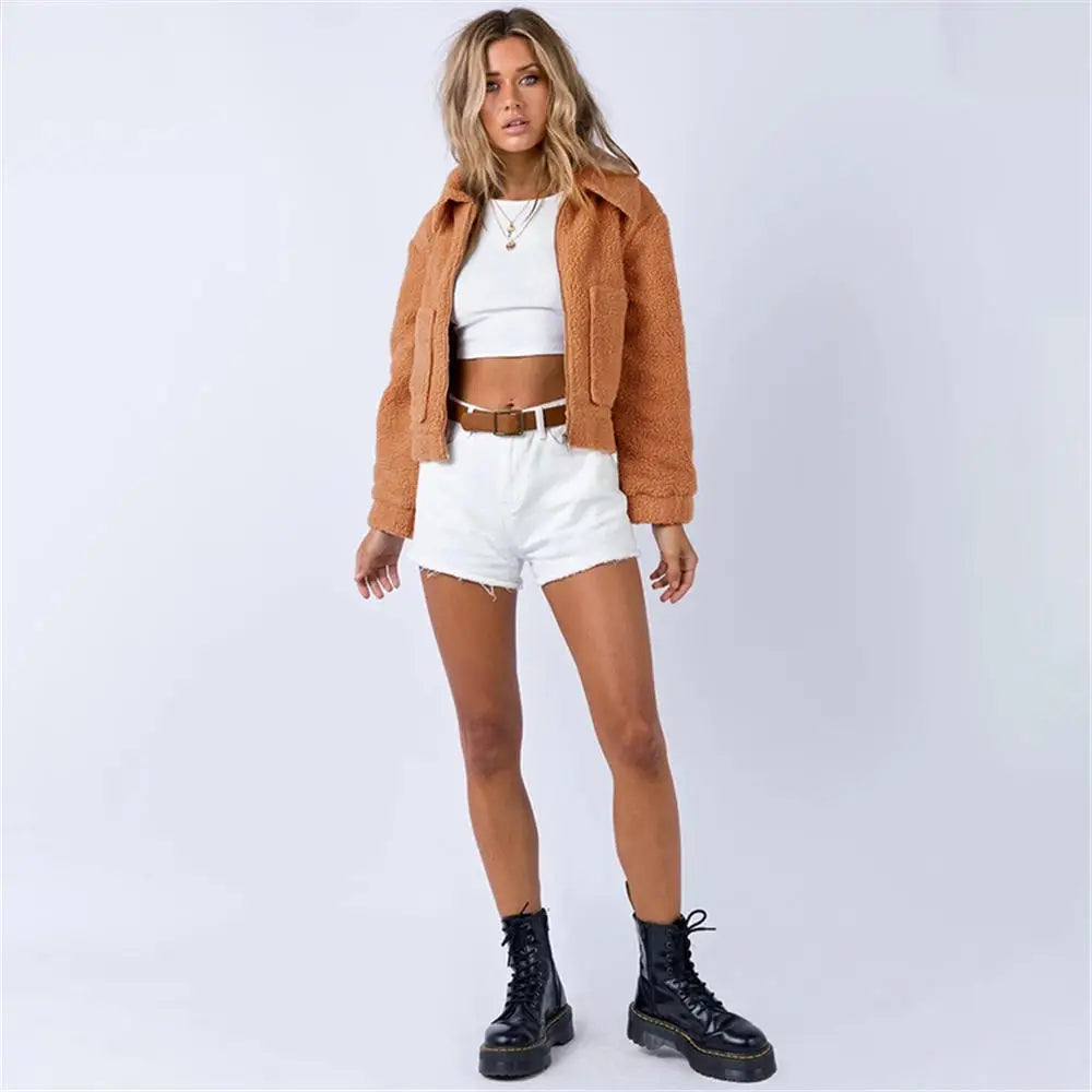 A woman stands against a plain light background wearing an all season tan sherpa fur cropped jacket, white crop top, white high-waisted shorts, a brown belt, and black lace-up boots. She has wavy hair and a relaxed pose.