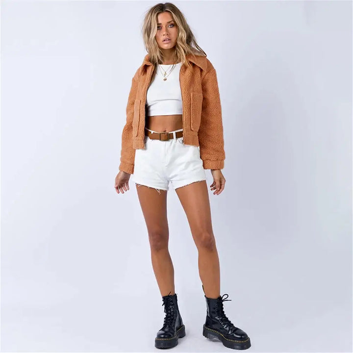 A woman stands against a plain light background wearing an all season tan sherpa fur cropped jacket, white crop top, white high-waisted shorts, a brown belt, and black lace-up boots. She has wavy hair and a relaxed pose.