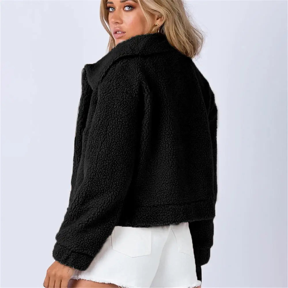 A woman wearing a black, sherpa fur teddy jacket and white denim shorts stands facing away, glancing over her shoulder against a plain light background.