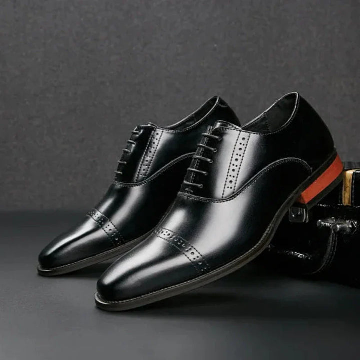 A pair of polished black Italian leather dress shoes with decorative stitching, cap toes, and a distinctive orange detail on the heel, featuring elegant hand-stitched detailing, displayed against a dark background.