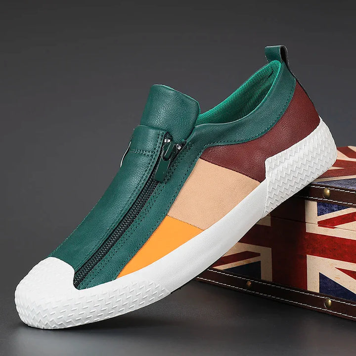 A stylish slip-on sneaker with a green leather front, orange and beige side panels, maroon heel, white cushioned sole, and zipper closure detail, displayed on a suitcase with a Union Jack design.