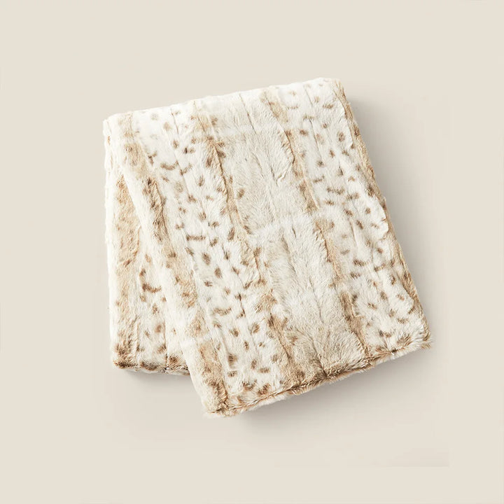 A folded ultra-soft luxurious blanket with a cream-colored base and light brown spotted pattern, reminiscent of a leopard print throw blanket, displayed on a plain beige background.