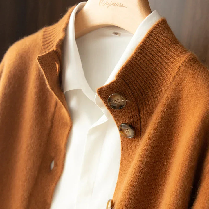 A close-up of a brown cashmere cardigan, crafted from 100% wool cashmere, worn over a white collared shirt on a wooden hanger. The cardigan features ribbed detailing on the collar, large buttons, and an ultra soft feel.
