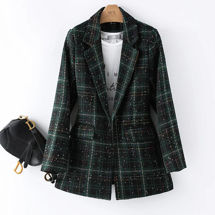 A green and black plaid formal blazer with subtle colored specks is hung on a hanger over a white printed t-shirt. A small black handbag with gold hardware is attached to the left sleeve, perfect for an office work suit look.