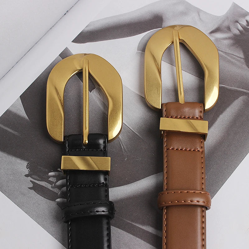Two genuine leather belts, one black and one brown, each with an oversized gold buckle, lie side by side on top of a grayscale fashion magazine featuring a woman in a dress.