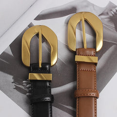 Two genuine leather belts, one black and one brown, each with an oversized gold buckle, lie side by side on top of a grayscale fashion magazine featuring a woman in a dress.