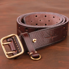 A coiled dark brown vintage leather belt with a textured finish, featuring a large brass buckle and multiple adjustment holes, placed on a wooden surface.