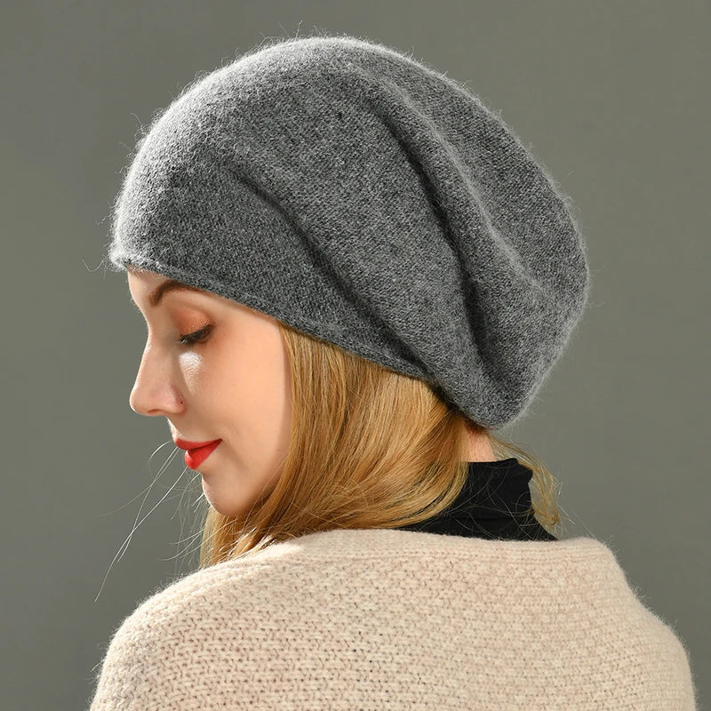 A woman with light brown hair wears a soft, oversized gray cashmere beret and a beige sweater. She is shown in profile against a plain gray background, looking downward with a calm expression.