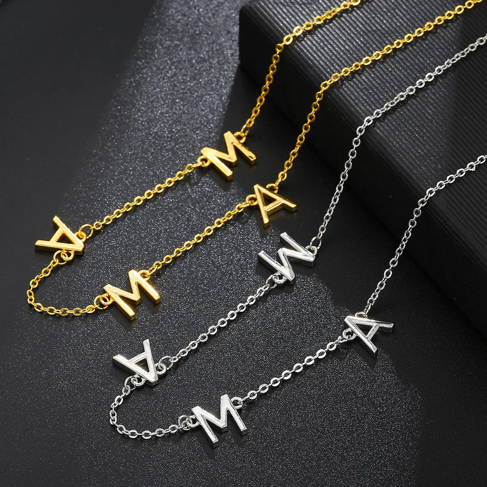 Two necklaces displayed on a black surface: one gold and one silver stainless steel necklace, each featuring three hanging letter charms A, M, and W. The minimalist jewelry chains are arranged neatly next to each other.