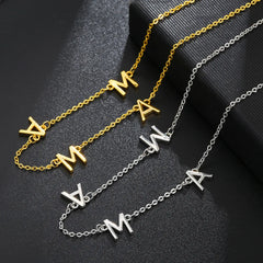 Two necklaces displayed on a black surface: one gold and one silver stainless steel necklace, each featuring three hanging letter charms A, M, and W. The minimalist jewelry chains are arranged neatly next to each other.