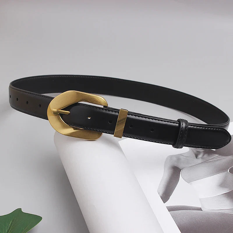 A genuine leather black belt with an oversized gold buckle is laid out on a magazine featuring a grayscale photo of a person. A green leaf is partially visible in the corner.