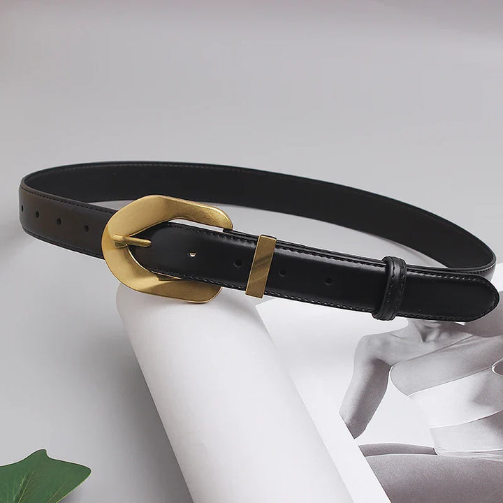 A genuine leather black belt with an oversized gold buckle is laid out on a magazine featuring a grayscale photo of a person. A green leaf is partially visible in the corner.