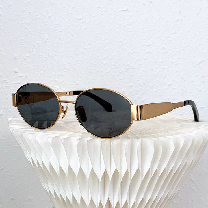 Vintage-inspired sunglasses with gold oval frames and dark UV400 protection lenses rest on a white, textured, geometric surface against a light-colored wall.