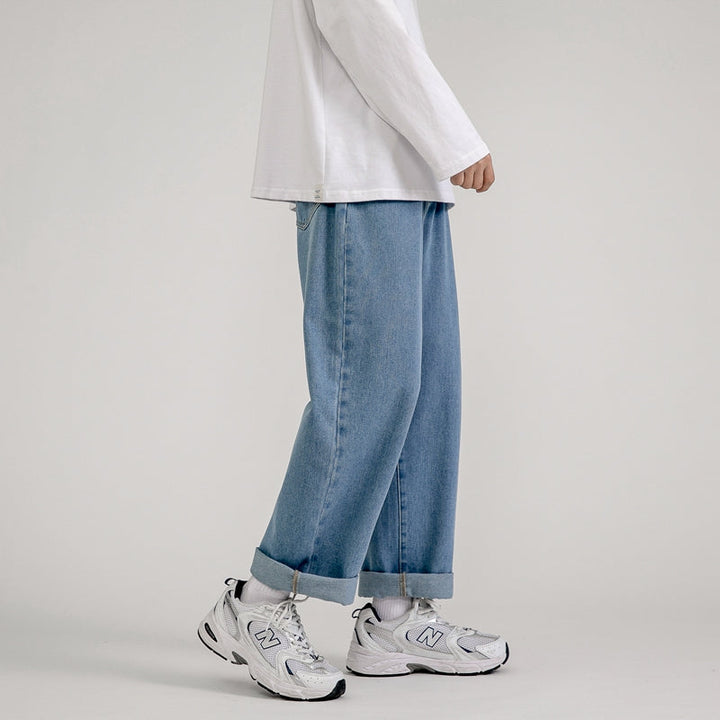 A person stands against a plain background, wearing a white long-sleeve shirt and Korean Fashion-inspired men’s baggy jeans with rolled cuffs, paired with white socks and New Balance sneakers.