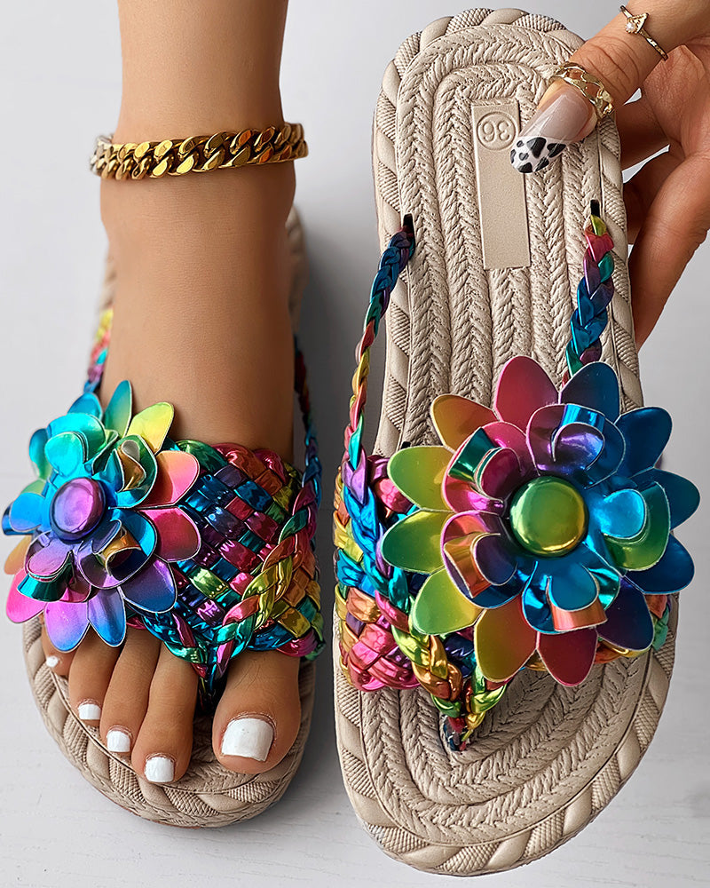A close-up of women's fashion casual shoes: braided beige sandals with large, colorful, metallic flower decorations. Perfect as beach flip flops or a vacation essential, they feature white toenail polish and a gold anklet while highlighting the details.