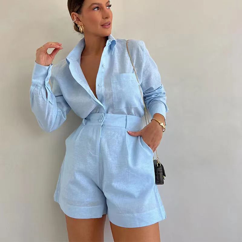 Woman wearing a light blue sustainable linen 2 piece set—long-sleeve button-up shirt tucked into matching high-waisted shorts—accessorized with a small crossbody bag, gold earrings, and bracelet, standing against a plain beige background.