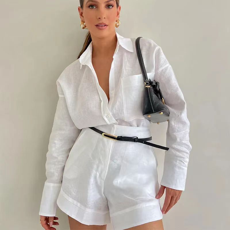 A woman wearing a white linen 2 piece set—button-down shirt tucked into high-waisted shorts—accessorized with a black belt, gold hoop earrings, and a small black shoulder bag, stands against a light-colored background.