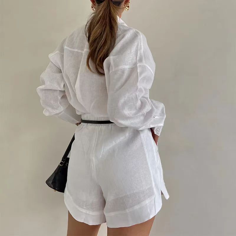 A woman with long brown hair in a ponytail wears a sheer white sustainable linen 2 piece set—long-sleeve shirt and matching shorts—accessorized with a black belt and shoulder bag, standing against a plain light-colored background.