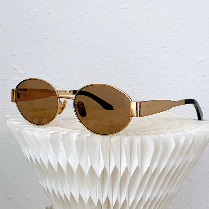 Gold-framed oval sunglasses with brown lenses rest on a white, textured, geometric surface against a plain white wall background, exuding Italian style and effortless sophistication.