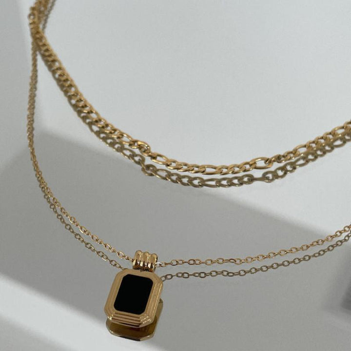 Two gold chain necklaces on a white surface; one necklace has a simple stainless steel chain, while the other features a rectangular black pendant set in gold. Shadows of the chains are visible in the background.