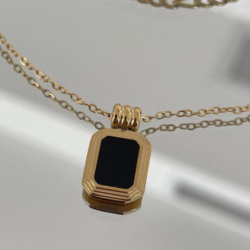 A close-up of a gold pendant necklace featuring a rectangular black centerpiece and crystal rhinestones, hanging from a delicate gold chain on a reflective surface.