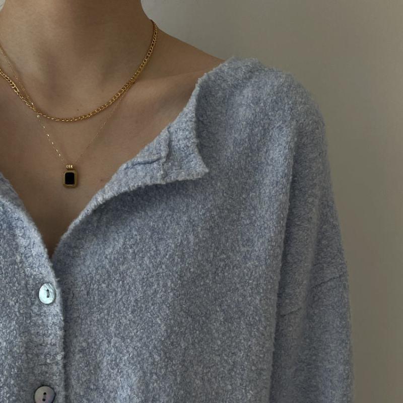 A person wearing a soft, textured light blue button-up shirt and two gold necklaces—one with a square pendant and the other a simple chain. The background is plain and neutral.