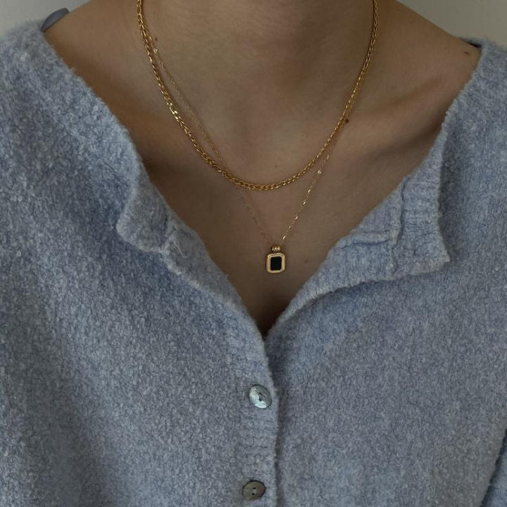 A person wearing a textured light blue button-up shirt and two layered gold necklaces, one featuring a square pendant. The image is cropped to show only the neck and upper chest area.