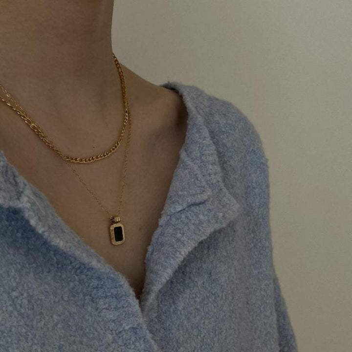 A person wearing a textured light blue top and two gold necklaces, one with a simple chain and the other featuring a rectangular black pendant accented by crystal rhinestone. The background is plain and neutral.