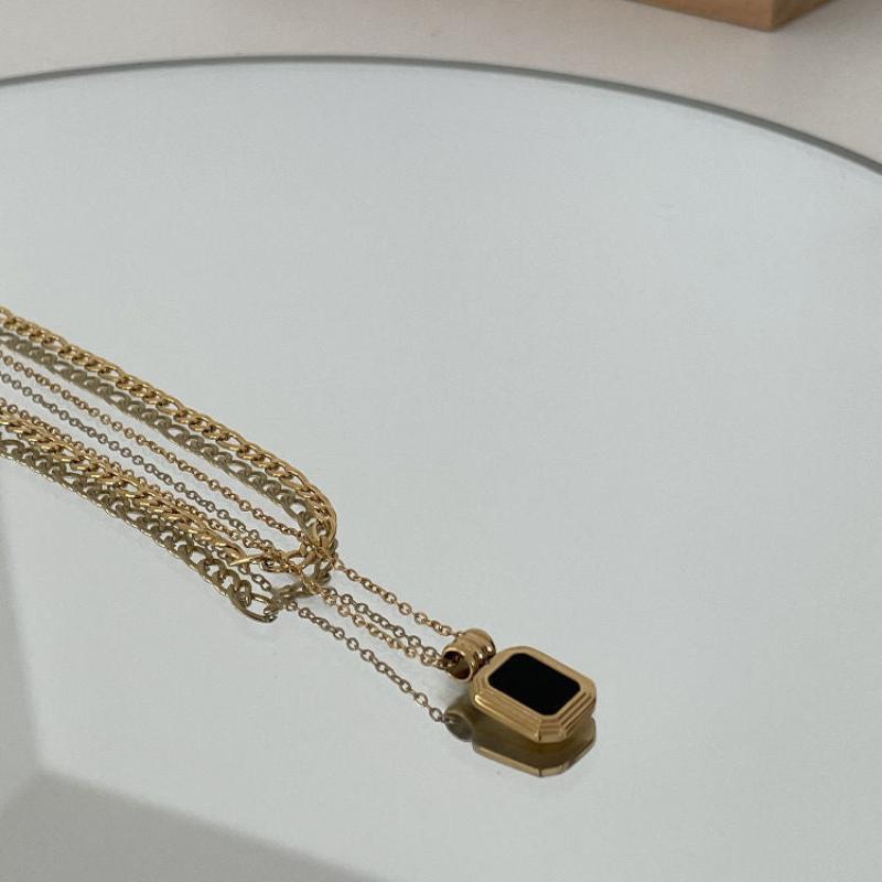 A gold chain necklace with a rectangular black pendant lies on a reflective surface, creating a subtle mirrored effect. The stainless steel finish adds durability and modern elegance to this striking piece.