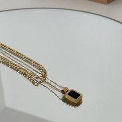 A gold chain necklace with a rectangular black pendant lies on a reflective surface, creating a subtle mirrored effect. The stainless steel finish adds durability and modern elegance to this striking piece.