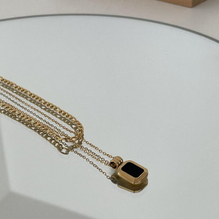 A gold chain necklace with a rectangular black pendant lies on a reflective surface, creating a subtle mirrored effect. The stainless steel finish adds durability and modern elegance to this striking piece.