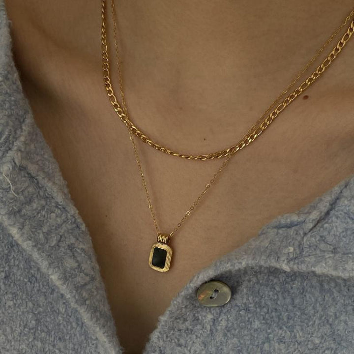 A close-up of a person wearing two gold necklaces; one is a simple chain and the other features a small, square pendant. The person is dressed in a light blue, textured button-up shirt.