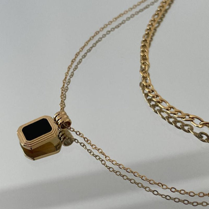 A gold necklace with a rectangular black gemstone pendant and a double chain design lies on a reflective surface alongside a second, thicker stainless steel gold chain.