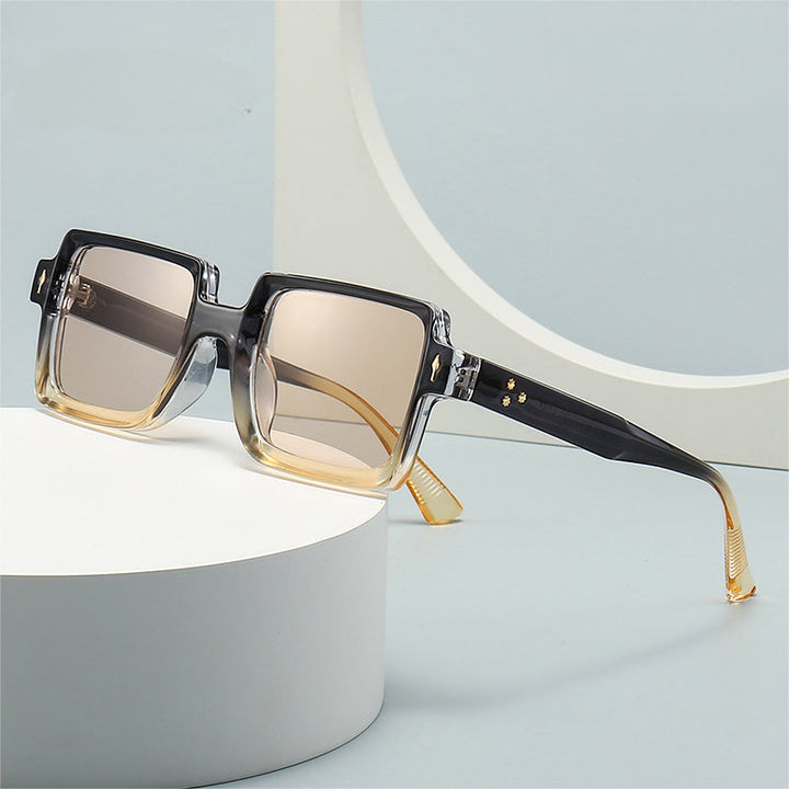 A pair of stylish, retro sunglasses with gradient gray-to-yellow frames and brown-tinted lenses is displayed on a white circular surface against a light blue background. These shades offer UV400 protection for your eyes.