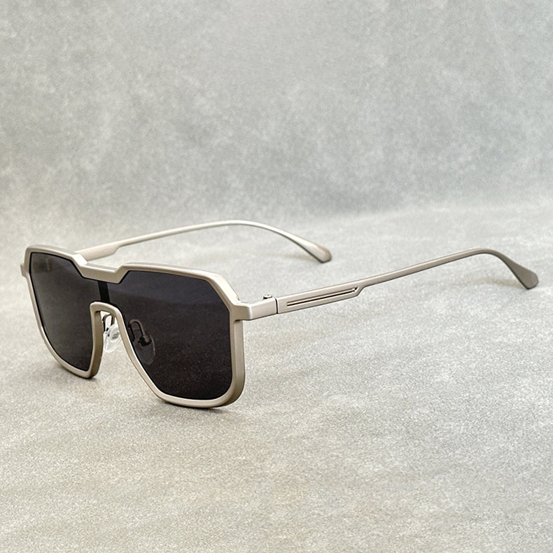 Silver-framed steampunk sunglasses with oversized, geometric black lenses are displayed on a light gray surface, their square frames and arms extended and angled slightly to the side.