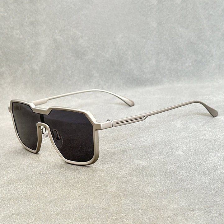 Silver-framed steampunk sunglasses with oversized, geometric black lenses are displayed on a light gray surface, their square frames and arms extended and angled slightly to the side.