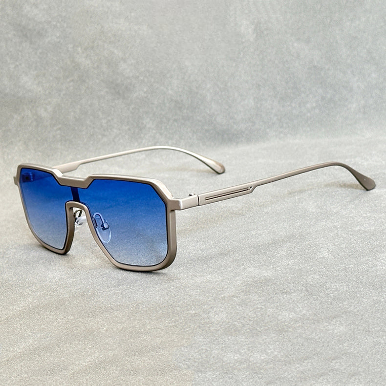 A pair of modern, geometric Heavy Metal Sunglasses with square metallic frames and blue gradient lenses rests on a light grey textured surface.