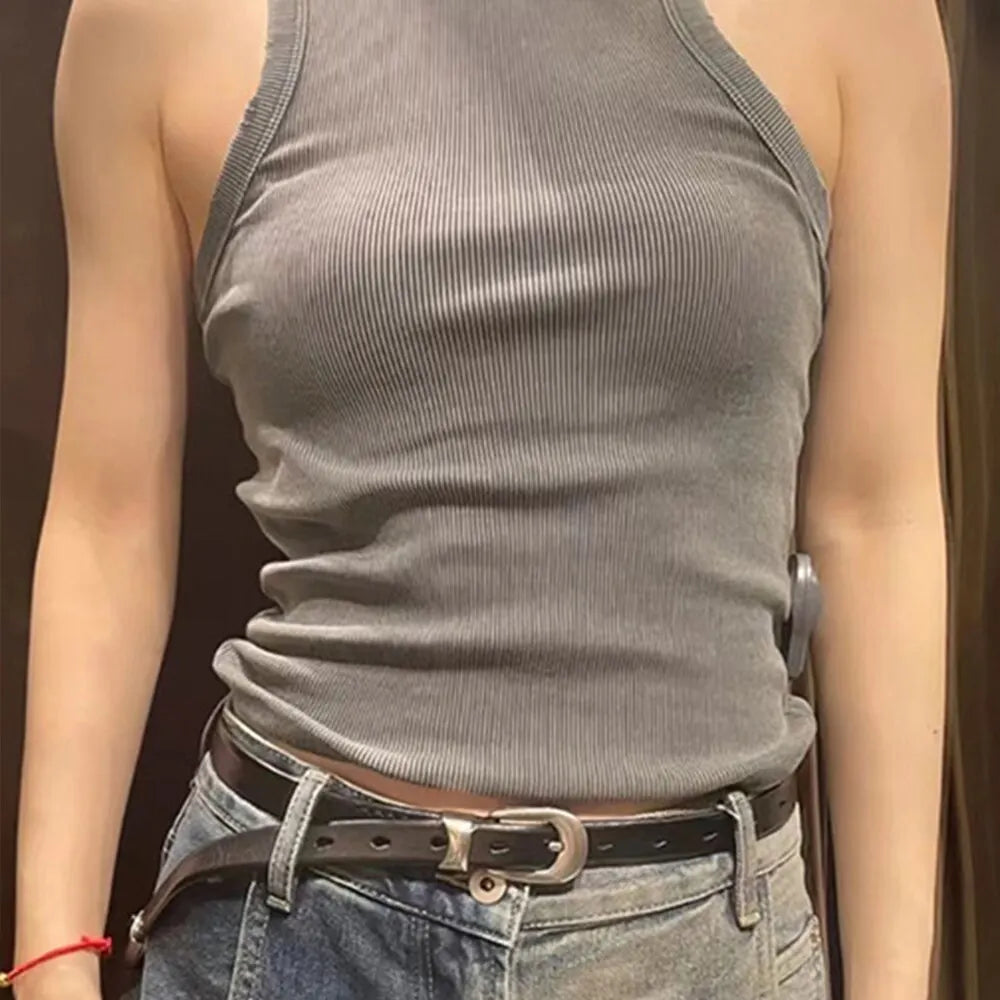 A person wearing a fitted, water wash rib women's tank top, a black belt, and blue jeans stands with arms by their sides. The background is out of focus and dark.