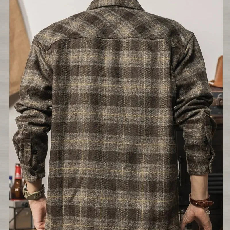 A person wearing a relaxed fit brown and gray plaid flannel shirt is seen from the back, arms slightly bent. The background features blurred objects like a bottle and a hat, adding to the cozy, layered look.