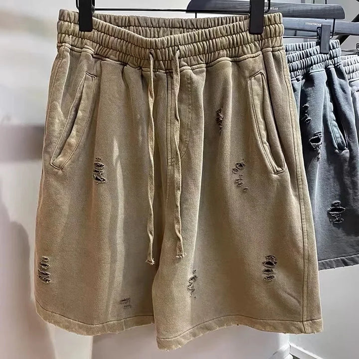 Beige vintage baggy shorts with an elastic waistband and cut-up design, featuring side pockets and a distressed look. Displayed hanging on a rack next to a similar grey pair.