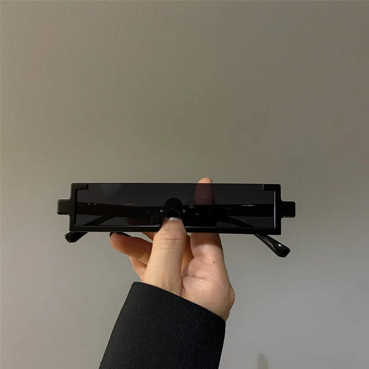 A hand with black-painted nails holds a pair of lightweight, rectangular black sunglasses with thick frames against a plain, light-colored background.