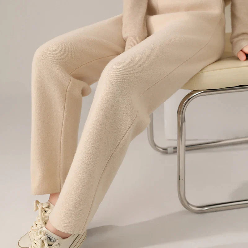 Person sitting on a light-colored chair, wearing cream-colored knit pants with a slim tailored leg, a matching sweater, and beige sneakers. Only the lower half of the body is visible against a neutral background.