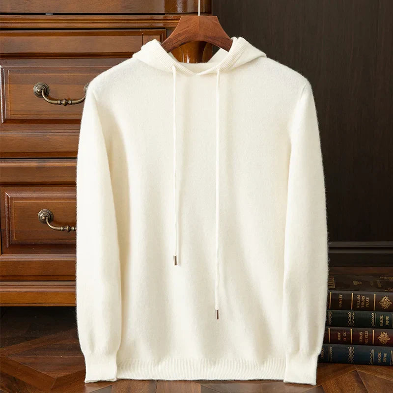A plain white layering sweater with drawstrings hangs on a wooden hanger in front of a wooden dresser and a stack of books on a wooden floor.