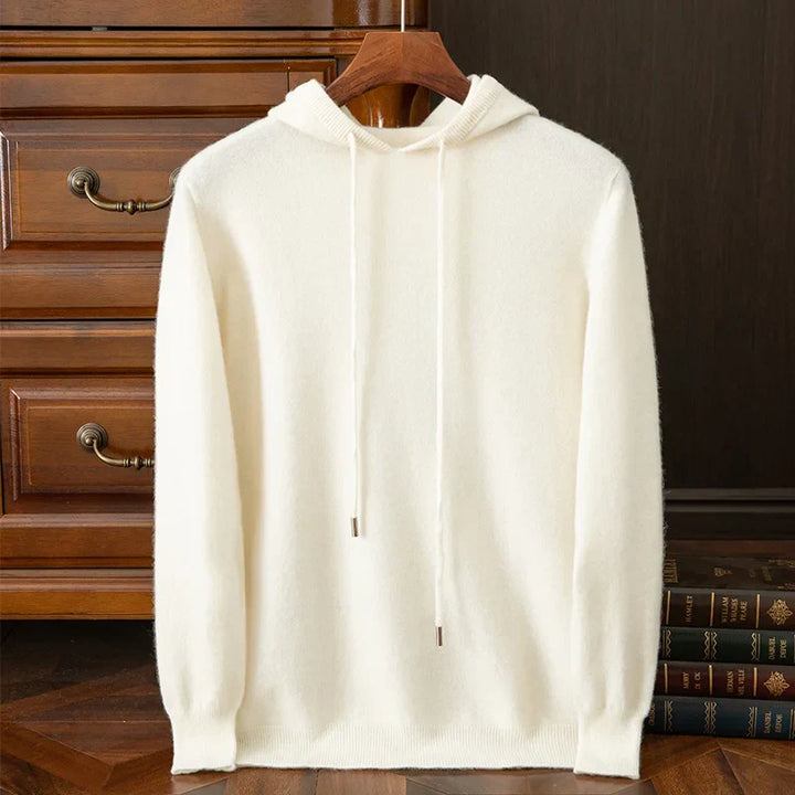 A plain white layering sweater with drawstrings hangs on a wooden hanger in front of a wooden dresser and a stack of books on a wooden floor.
