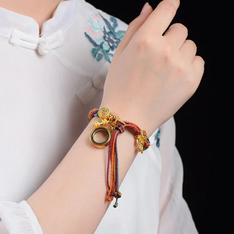 A person wearing a white, floral-embroidered blouse showcases an adjustable, colorful braided bracelet with gold charms, a ring-shaped pendant, and Tibetan craftsmanship on their wrist.