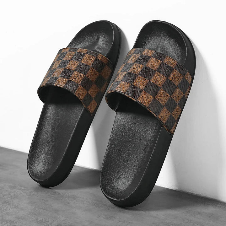 A pair of black streetwear slides with brown and black checkerboard patterned straps, placed on a gray floor against a white wall.