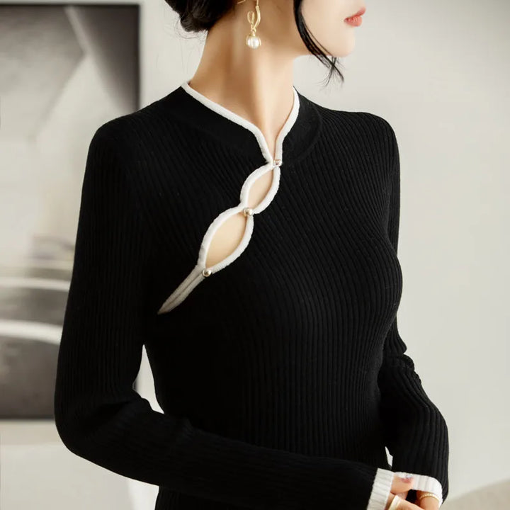 A woman wearing a black ribbed knit long-sleeve dress with two keyhole designs near the neckline, outlined in white trim and fastened with pearl-like buttons. She has pearl drop earrings and her hair is styled up.