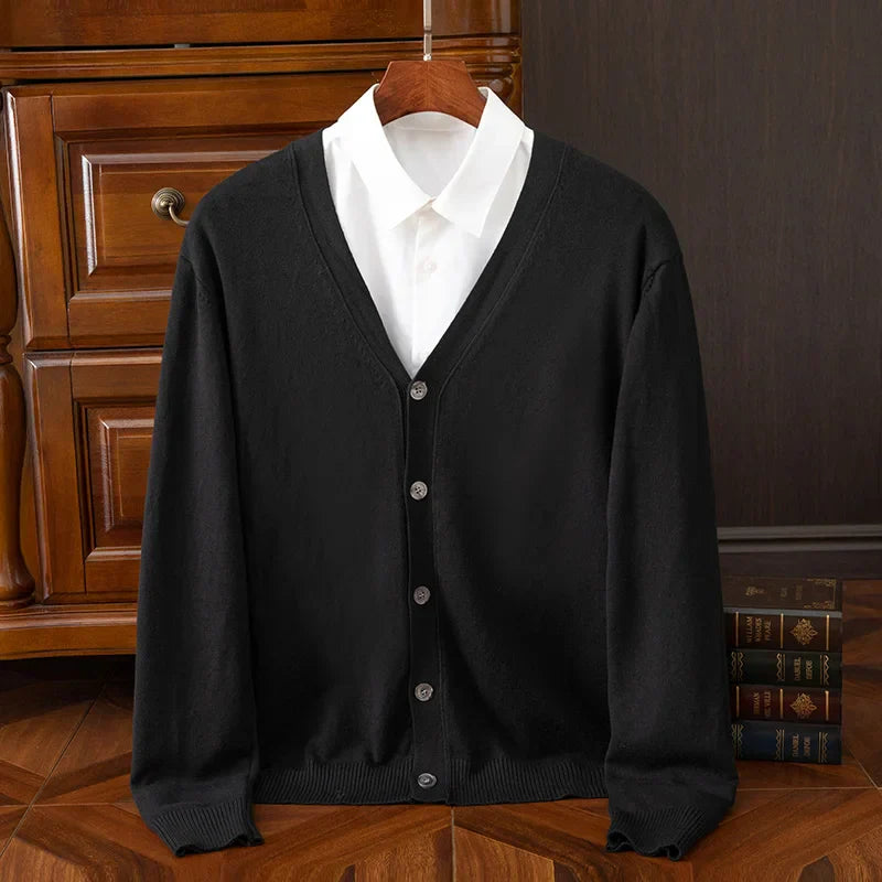 A black cashmere wool blend button-up cardigan with a V-neckline is layered over a white collared shirt, displayed on a wooden hanger in front of a wooden drawer and some stacked books on a wooden floor.